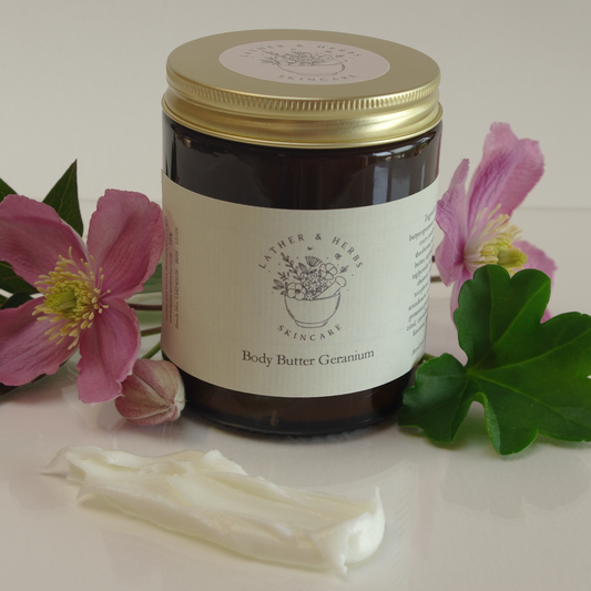 Amber glass jar with gold aluminium lid and flowers around with geranium scented body butter 